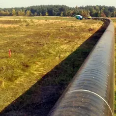 Długi, czarny rurociąg „Przyjaźń” wijący się przez łąkę w kierunku lasu. W oddali widać maszyny budowlane.