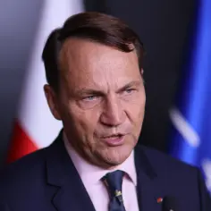 Minister spraw zagranicznych w garniturze, z polską flagą i flagą NATO w tle.