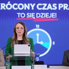 Kobieta przemawia na tle hasła "SKRÓCONY CZAS PRACY TO SIĘ DZIEJE!". Obok niej zegar symbolizujący skrócenie czasu pracy.