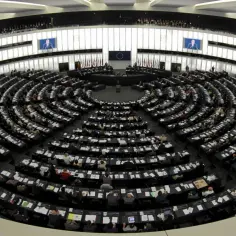 Wielka sala plenarna, gdzie obraduje **parlament europejski**. Posłowie siedzą w półkolistych rzędach, a na ekranach wyświetlane są przemówienia.
