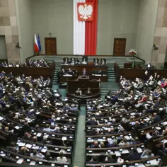Wielka sala obrad, gdzie odbywa się **zgromadzenie narodowe**. Posłowie siedzą w ławach, a na mównicy przemawia jeden z nich.