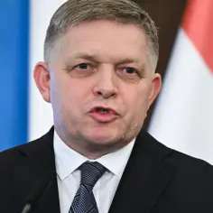 Premier Słowacji w garniturze i krawacie, przemawia na tle flag.