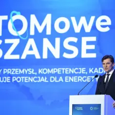 Mężczyzna w garniturze przemawia na tle napisu "ATOMOWE SZANSE", podkreślając rozwój polskiego przemysłu i kompetencji dla energetyki jądrowej.