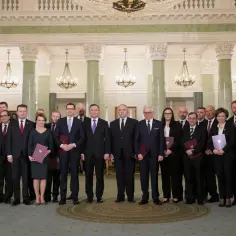 Grupa osób w eleganckich strojach, prawdopodobnie przedstawiciele ministerstw w Polsce, pozuje do zdjęcia w przestronnym, ozdobnym wnętrzu.