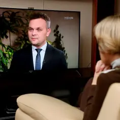 Prezes TVP w garniturze udziela wywiadu. Kobieta ogląda go z kanapy.