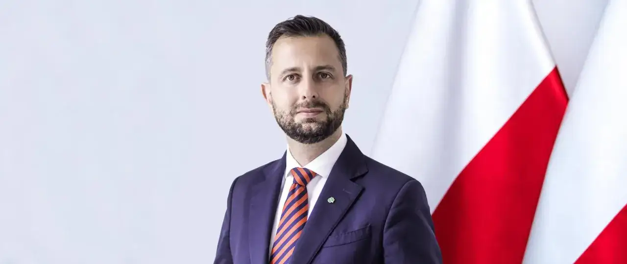 Mężczyzna w garniturze, z brodą, obok polskiej flagi. To minister obrony, gotowy do działania.