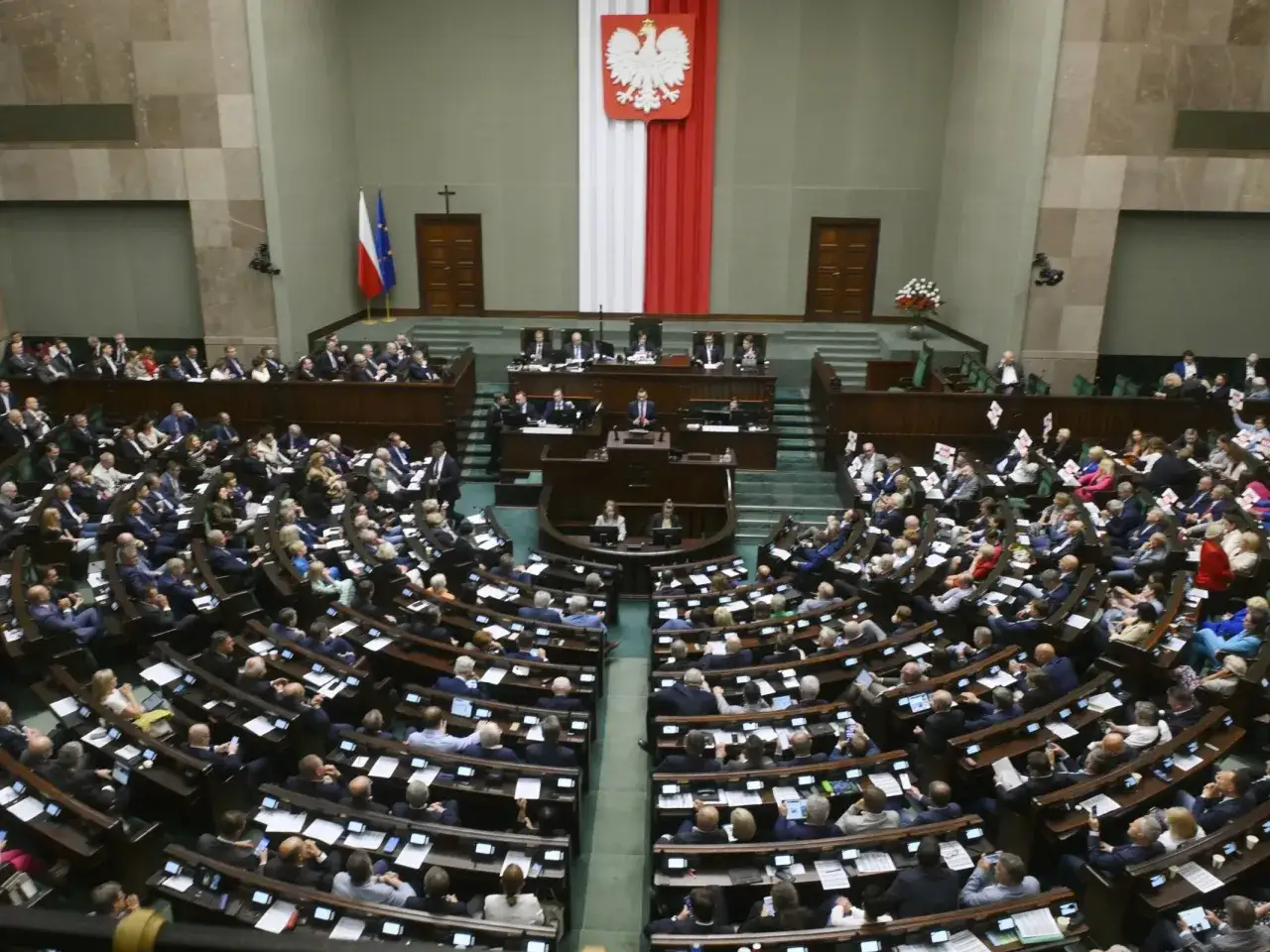 Wielkie zgromadzenie narodowe w Polsce. Posłowie siedzą w ławach, a na mównicy przemawia polityk. Nad nimi dumnie powiewa polska flaga.