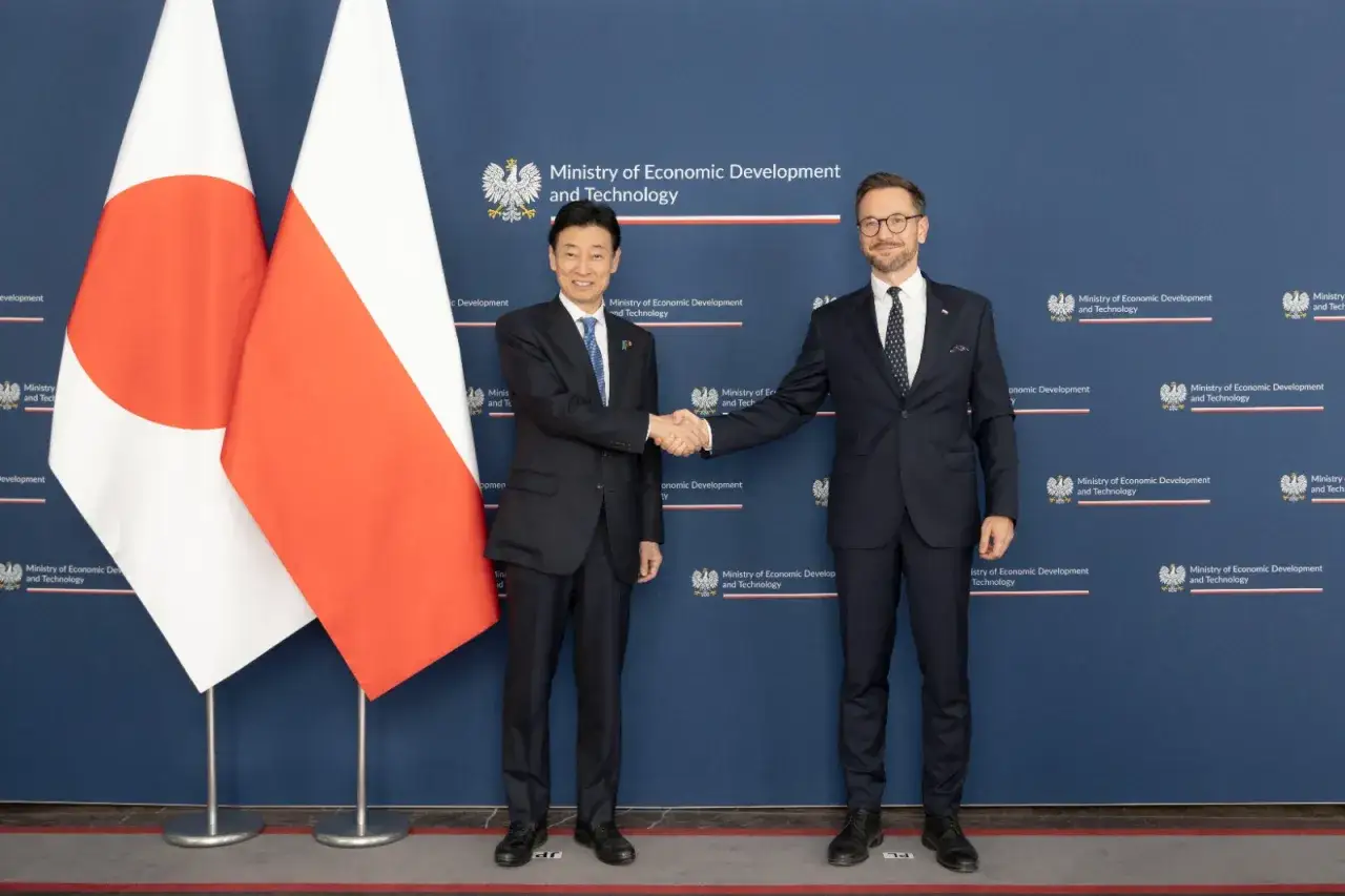 Dwaj mężczyźni ściskają sobie dłonie na tle flag Polski i Japonii. Współpraca polsko-japońska kwitnie.